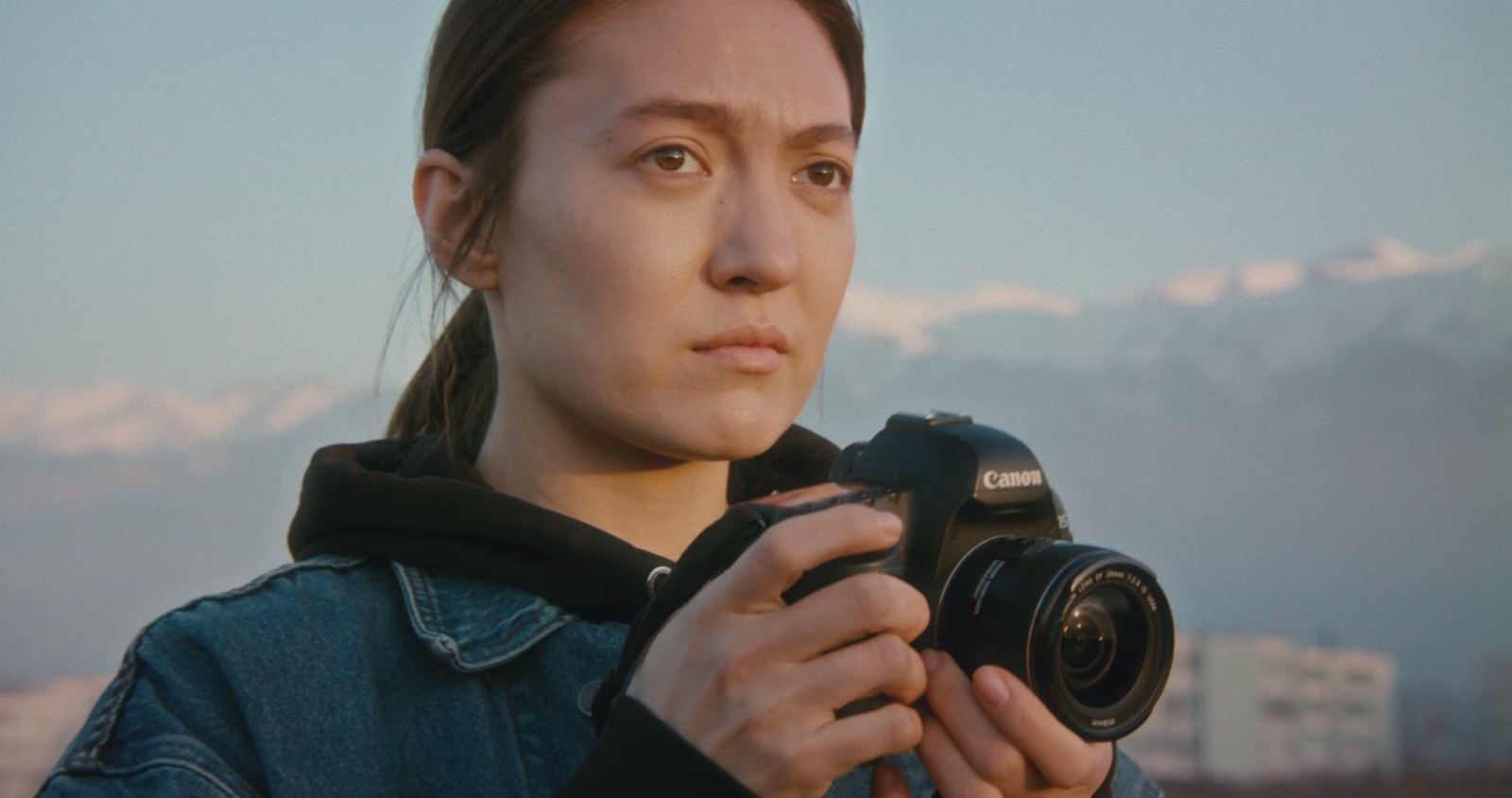 Merey stands outdoors at dusk holding her camera, snow-capped mountains soft behind her.