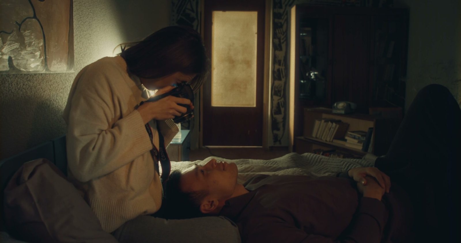 Merey leans over a man lying on a bed, holding her camera close to his face while he looks up at her.