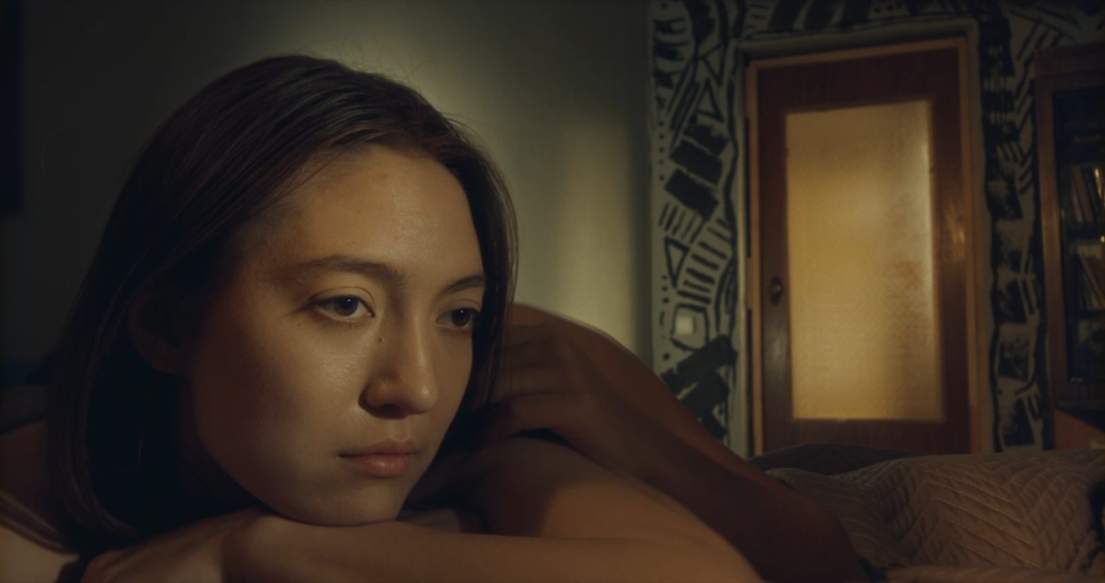 Merey lies on a bed, her head resting on her arms, looking away into the dim room.