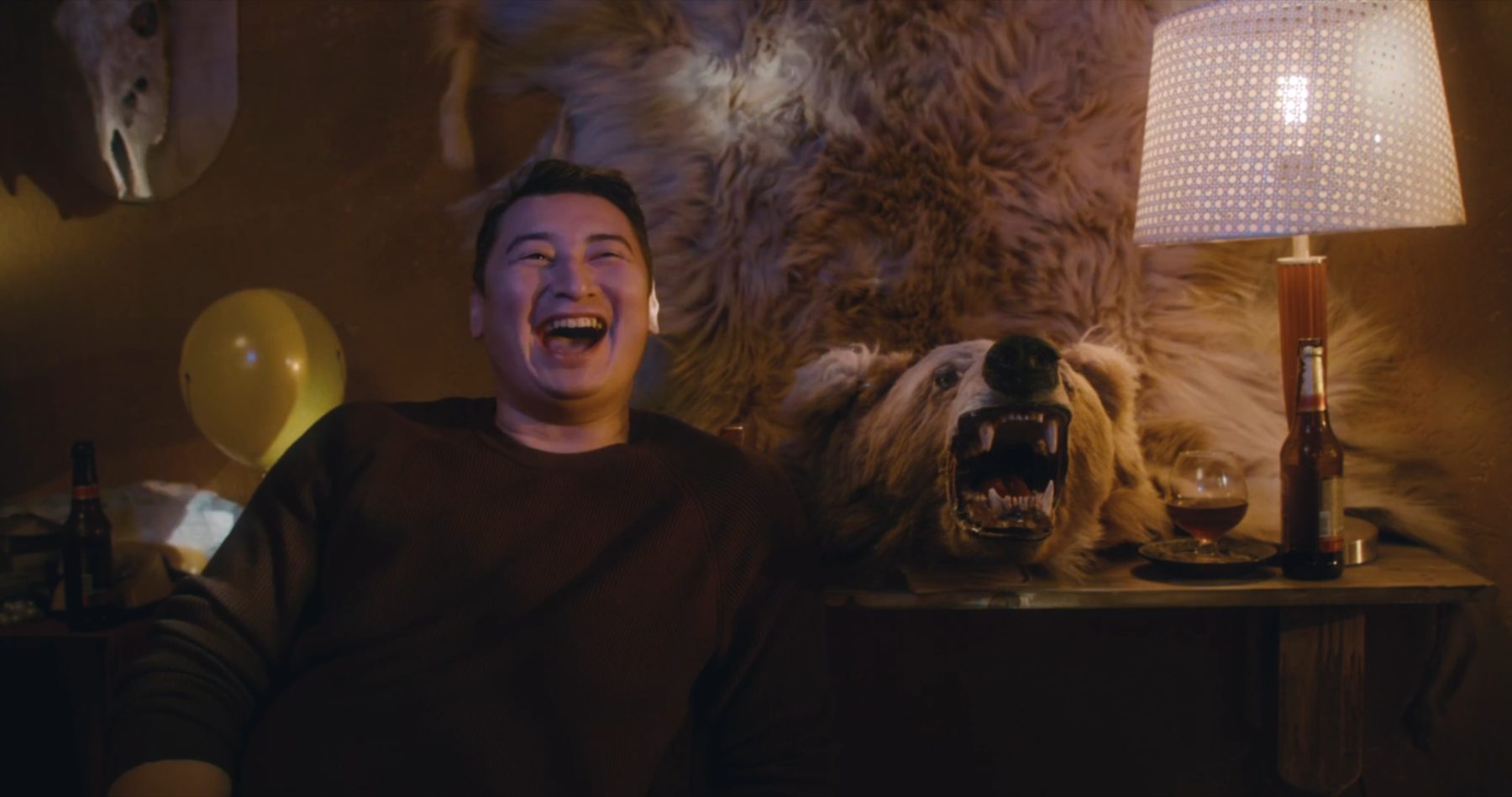 A man laughs open-mouthed on a sofa, a party balloon beside him and a snarling bear-skin rug pinned to the wall behind his head.